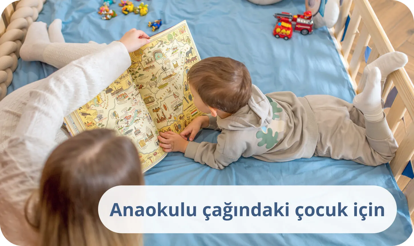 Annesiyle birlikte çam ağacından bir yatakta, mavi nevresimler üzerinde renkli bir kitap okuyan küçük çocuk.